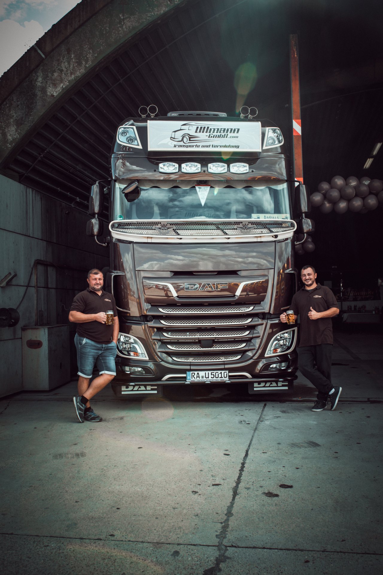 Zwei Männer mit Biergläsern stehen vor einem braunen DAF-LKW; oben am LKW steht "Illmann GmbH".